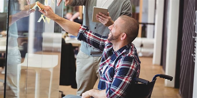 Ein Mann im Rollstuhl skizziert im Büro eine Idee mit seinem Arbeitskollegen an einer Glaswand. Ein Mann im Rollstuhl skizziert im Büro eine Idee mit seinem Arbeitskollegen an einer Glaswand.