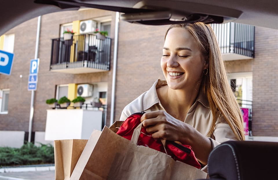 Eine Frau lächelt, während sie eine Tüte aus einem Auto nimmt, mit Wohnungen im Hintergrund. Eine Frau lächelt, während sie eine Tüte aus einem Auto nimmt, mit Wohnungen im Hintergrund.