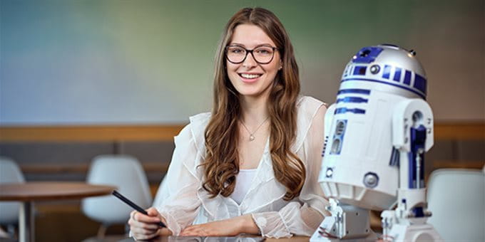 Eine Person sitzt an einem Tisch in einem modernen Raum und arbeitet mit einem Tablet und Stift. Neben ihr steht ein Modell des Roboters R2-D2 aus Star Wars. Eine Person sitzt an einem Tisch in einem modernen Raum und arbeitet mit einem Tablet und Stift. Neben ihr steht ein Modell des Roboters R2-D2 aus Star Wars.