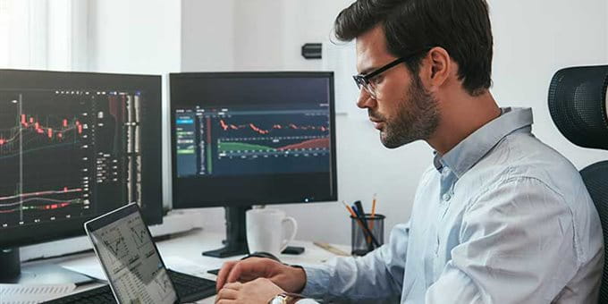 A man with glasses works on a laptop in front of two monitors displaying graphs. A man with glasses works on a laptop in front of two monitors displaying graphs.