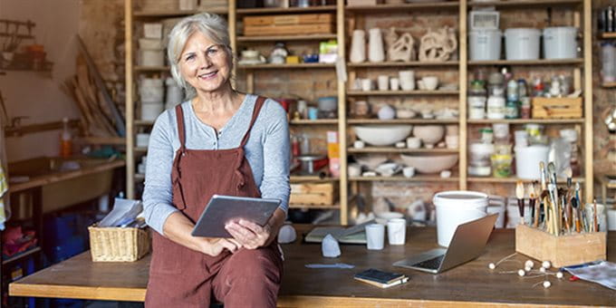 An older woman smiles while sitting at a table in an art studio, holding a tablet. An older woman smiles while sitting at a table in an art studio, holding a tablet.