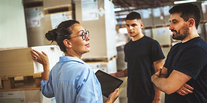 Three people in a warehouse are talking. A woman is showing a tablet while two men listen. Three people in a warehouse are talking. A woman is showing a tablet while two men listen.