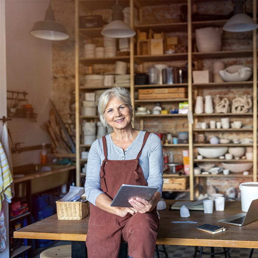 An older woman sits with a tablet on a table in a creative workspace. An older woman sits with a tablet on a table in a creative workspace.