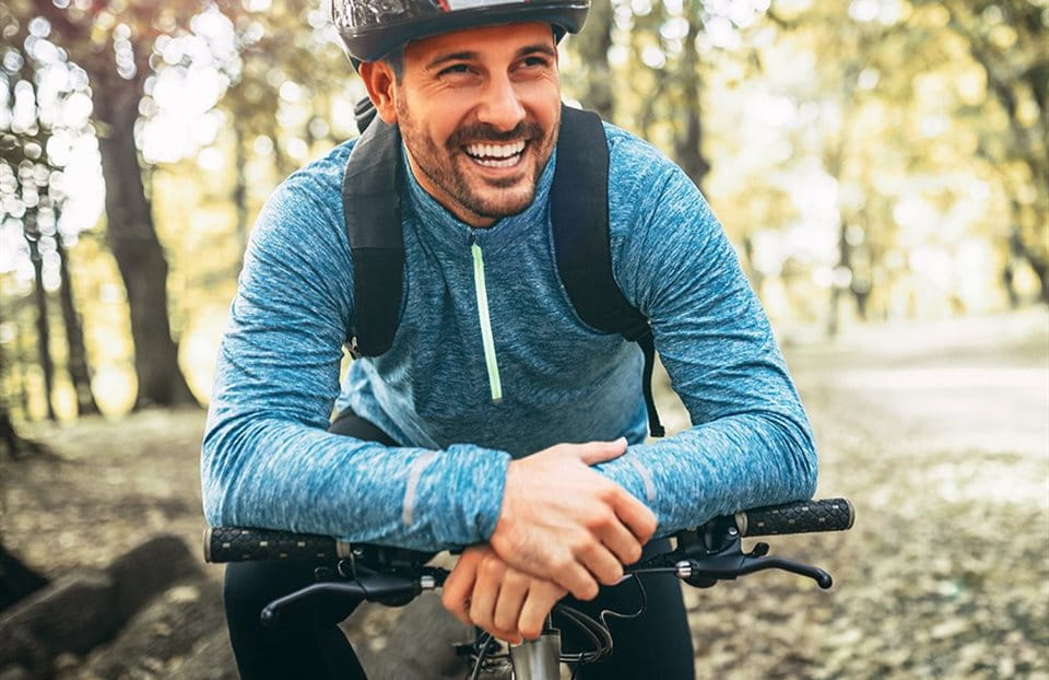 A smiling man in a blue shirt and helmet on a bike in a forest. A smiling man in a blue shirt and helmet on a bike in a forest.