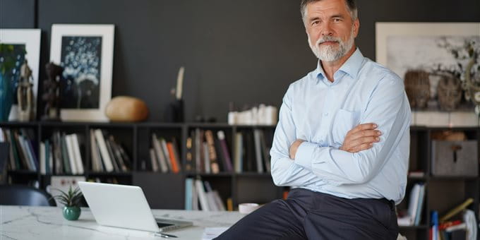 A man in light clothing sits with his arms crossed on a desk in a modern office with shelves, books, and a laptop. A man in light clothing sits with his arms crossed on a desk in a modern office with shelves, books, and a laptop.