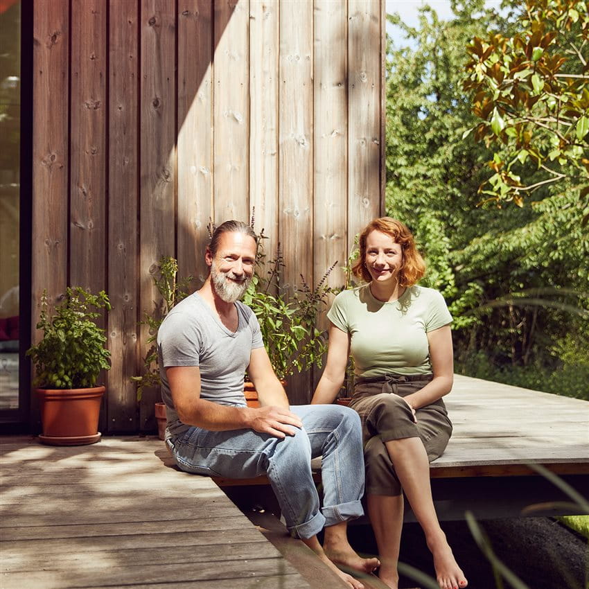Un couple est assis pieds nus sur une terrasse en bois devant une maison moderne en bois, entourée de plantes. Un couple est assis pieds nus sur une terrasse en bois devant une maison moderne en bois, entourée de plantes.
