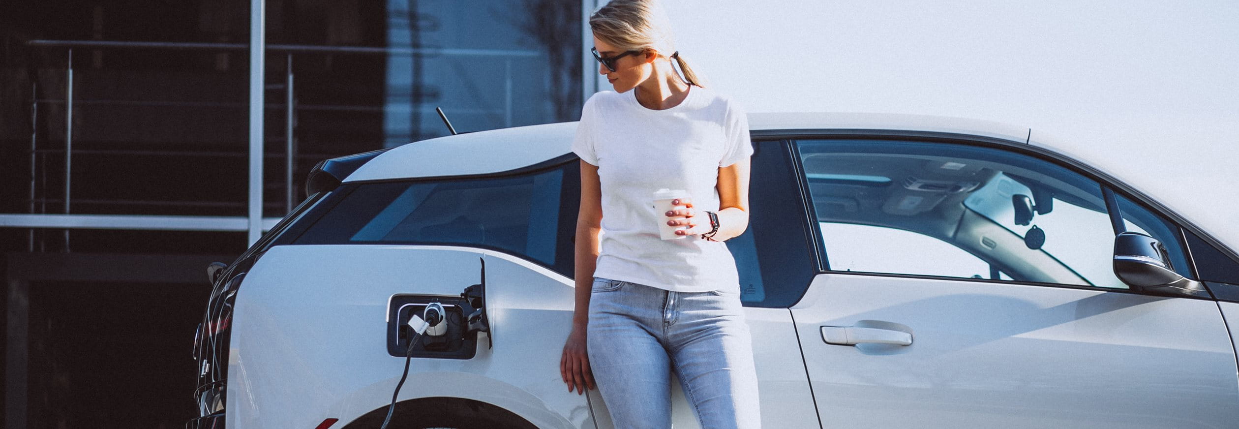 Woman stands in front of her car at the electric charging station and drinks coffee  Woman stands in front of her car at the electric charging station and drinks coffee