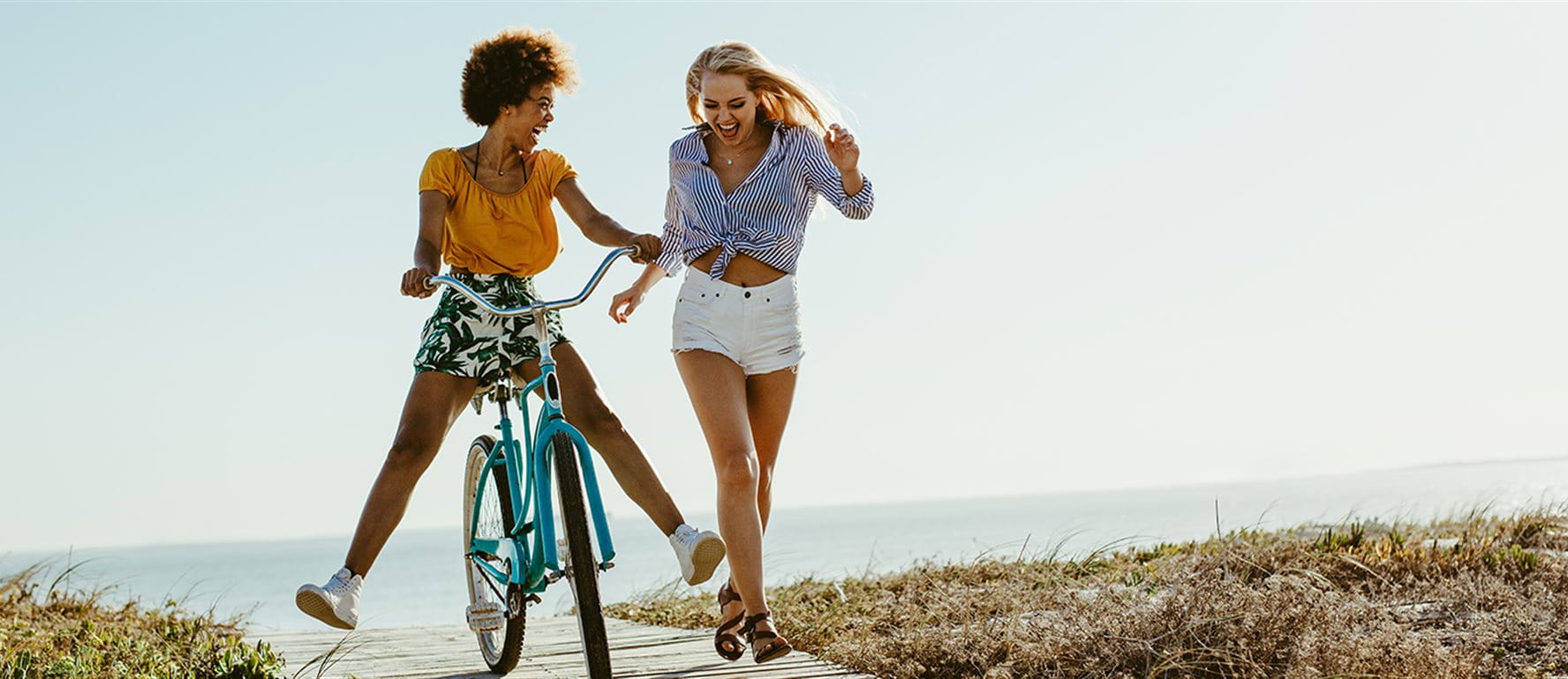 Two young women at the beach – one is riding a bike and smiling, the other is walking alongside. Two young women at the beach – one is riding a bike and smiling, the other is walking alongside.