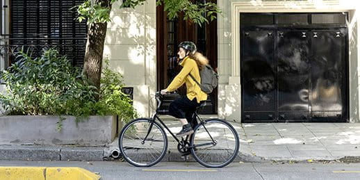 A person on a bicycle rides along the street, with greenery and a building in the background. A person on a bicycle rides along the street, with greenery and a building in the background.
