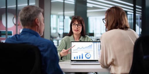 Three people are discussing in a modern office. One person points at a graph on a laptop. Three people are discussing in a modern office. One person points at a graph on a laptop.