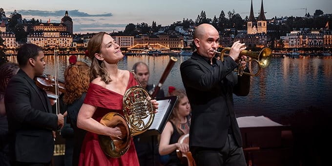 Open-air concert with musicians in front of a lake and city backdrop. Open-air concert with musicians in front of a lake and city backdrop.