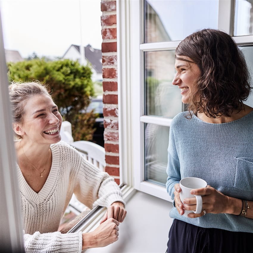 Zwei Frauen unterhalten sich entspannt am offenen Fenster eines Hauses. Eine Frau steht draußen und lehnt sich auf das Fensterbrett, die andere steht drinnen und hält eine Tasse Kaffee. Im Hintergrund sind ein Garten und Nachbarhäuser zu sehen. Zwei Frauen unterhalten sich entspannt am offenen Fenster eines Hauses. Eine Frau steht draußen und lehnt sich auf das Fensterbrett, die andere steht drinnen und hält eine Tasse Kaffee. Im Hintergrund sind ein Garten und Nachbarhäuser zu sehen.