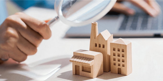 A hand holds a magnifying glass over wooden miniature buildings on a table. A hand holds a magnifying glass over wooden miniature buildings on a table.